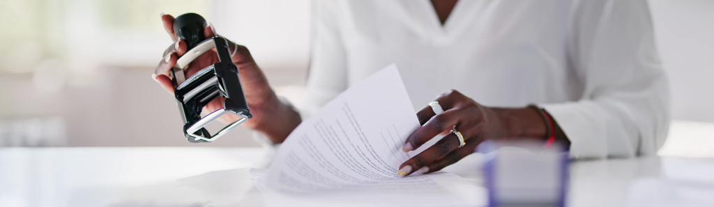 Close-up of hands holding an official notary stamp above legal documents on a white desk, demonstrating the document certification process.