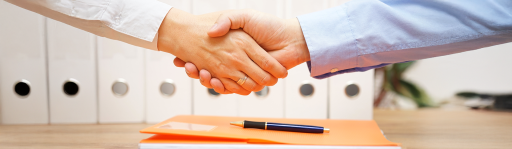 Professional handshake over a desk with an orange folder and pen, with white file folders visible in the background, symbolizing agreement on legal documents.